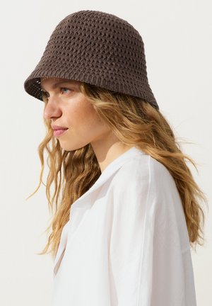 Tânără cu păr blond ondulat, purtând o pălărie maro croșetată de tip bucket și o cămașă albă, orientată spre stânga pe un fundal simplu.
