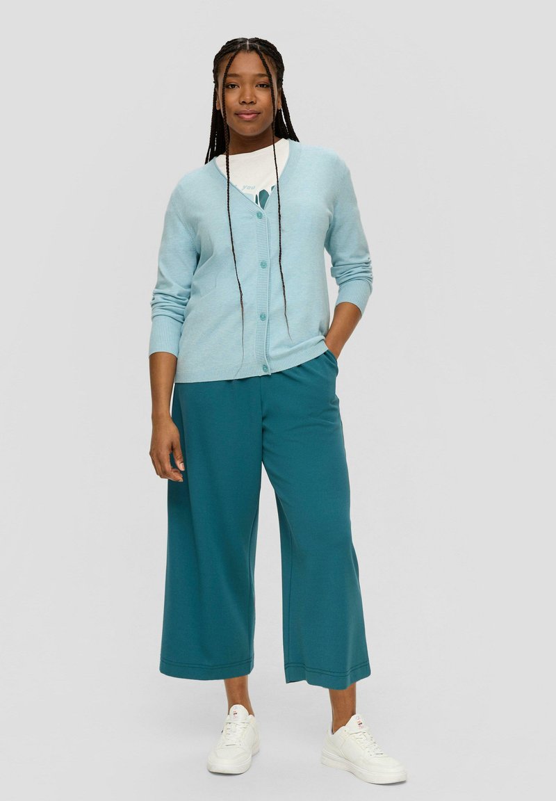 Cardigan bleu clair avec un devant à bouton, associé à un pantalon large teal et des baskets blanches. Texture lisse avec des accents côtelés.