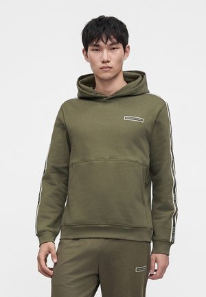 Mladý muž nosí olivovozelenú mikinu s kapucňou a zodpovedajúce tepláky s logom EA7 Emporio Armani na hrudi, rukávoch a nohaviciach.