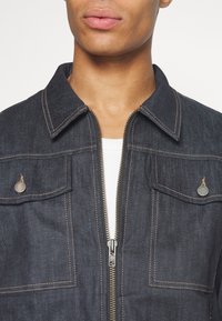 Primer plano de una persona que lleva una chaqueta de mezclilla oscura con costuras beige, bolsillos a presión de metal y una parte delantera parcialmente cerrada sobre una camisa blanca.