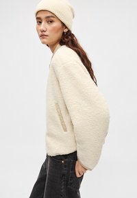 Μπλούζα από μπεζ φλις με υφή, φαρδιά μανίκια και πλαϊνή τσέπη, σε συνδυασμό με σκούρο τζιν και ανοιχτό σκουφάκι.