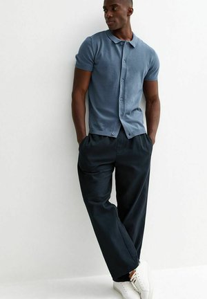 Uomo appoggiato al muro con le mani nelle tasche, indossa una camicia blu a maniche corte abbottonata, pantaloni neri e sneakers bianche.