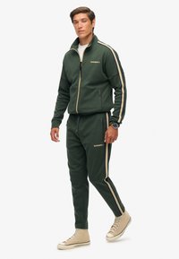 Mørkegrøn træningsdragt med beige sidelister, bestående af en zip-up jakke og bukser med elastisk talje. Skoene er lys beige med snørebånd.