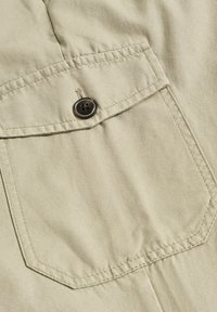 Helles beige Gewebe mit strukturierter Oberfläche. Verfügt über eine Brusttasche mit einem braunen Knopf und verstärkten Nähten entlang der Kanten.