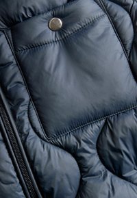 Närbild av mörkblå quiltad jacktyg med synlig ficka, metalltryckknapp och dragkedjesöm.