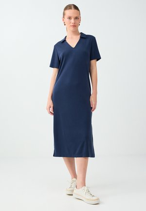 Femme debout portant une robe midi bleu marine à manches courtes avec col et des baskets blanches sur un fond clair uni.