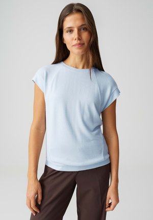 Giovane donna con capelli castani lunghi che indossa una maglia a maniche corte azzurra e pantaloni marrone scuro, in piedi davanti a uno sfondo neutro.