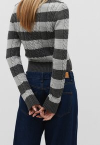 Pull court en tricot à rayures gris et gris foncé, avec une texture en tricot à cablé et des poignets côtelés, associé à un jean large bleu foncé.