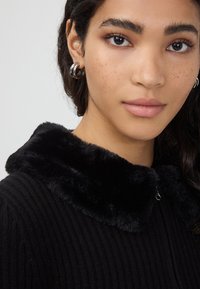 Czarna, żeberkowana odzież z miękkim, pluszowym kołnierzem. Zbliżenie na twarz osoby, pokazująca kolczyki w kształcie kół oraz naturalną cerę.