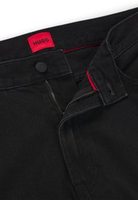 Svarte denimbukser med en rød HUGO-lapp. Egenskaper inkluderer glidelås, knappelukking og synlig søm. Glatt tekstur og tettsittende design.