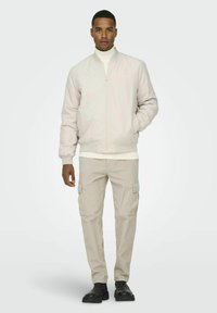 Ljusbeige bomberjacka med ribbade ärmar, kombinerad med en krämfärgad turtleneck och beige cargo-byxor med sidofickor. Svarta ankelskurna stövlar.