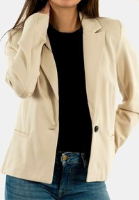 Beige blazer with a tailored fit, single button closure, two front pockets, and smooth fabric texture. Worn over a black top.
