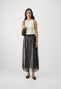 Top senza maniche color crema con scollo rotondo, abbinato a una lunga gonna nera trasparente e plissettata. La modella tiene in mano una piccola borsa nera e indossa delle scarpe basse.