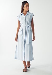 Donna che indossa un vestito midi senza maniche di colore azzurro chiaro con cintura e dettagli a bottoni, in piedi contro uno sfondo bianco uniforme.