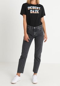 Černé tričko s nápisem "DESERT DAZE" v pestrých barvách, doladěné s vysokými šedými džíny a bílými teniskami. Klasický outfit.