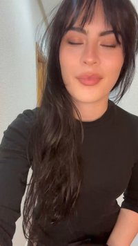 Jeune femme aux longs cheveux foncés et à la frange, portant un haut noir, les yeux fermés dans un intérieur à la lumière douce.