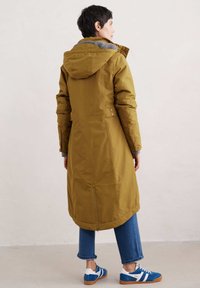 Manteau long vert olive avec une capuche, doté d'une doublure grise, d'accents de boutons sur les manches et d'un ourlet arrière plus long, associé à un denim bleu.