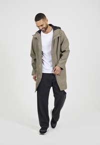 Giacca impermeabile color khaki con cappuccio, bottoni e tasche, indossata sopra una camicia bianca e pantaloni neri, con una vestibilità comoda e una superficie liscia.