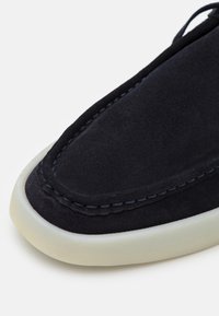Navy suede slip-on shoe featuring white rubber sole, contrast stitching, and smooth, matte texture. Rounded toe with minimalistic design.
