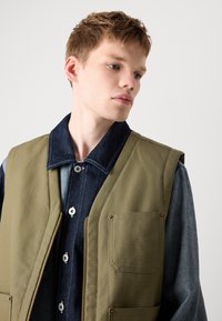 Olive green sleeveless vest with a zip front, two patch pockets, and a denim shirt underneath featuring a dark collar and buttons.