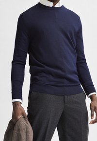 Mörkblå stickad tröja med rund halsringning och ribbad fåll, bärs över en vit skjorta med krage, tillsammans med grå byxor.