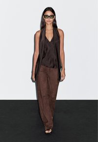 Top marrone senza maniche con scollatura a V e design drappeggiato, abbinato a pantaloni larghi marroni in una morbida texture, completato da occhiali da sole verdi.