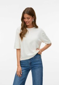 Kortærmet, cremefarvet top lavet af blødt stof med en rund halsudskæring. Kombination med blå denimjeans. Ingen synlige mønstre eller dekorative elementer.