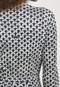 Schwarzes und weißes, gemustertes Kleidungsstück mit einem sich wiederholenden Fächerdesign. Das Material wirkt weich, mit einer strukturierten Oberfläche und einem sichtbaren Markenlabel.