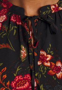 Schwarze Blumenbluse mit gerafftem Ausschnitt, die rote und orangefarbene Blumen sowie grüne Blätter zeigt. Mit einem kleinen Knotenverschluss vorne.