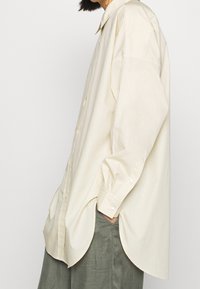 Chemise beige clair boutonnée avec une coupe décontractée, un ourlet arrière plus long et un col classique. Caractéristiques manches longues avec poignets à boutons et texture lisse.