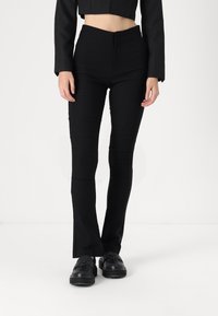 Calças pretas ajustadas com cintura alta, apresentando uma textura suave e costuras mínimas, acompanhadas de sapatos pretos. Cor sólida, sem padrões.