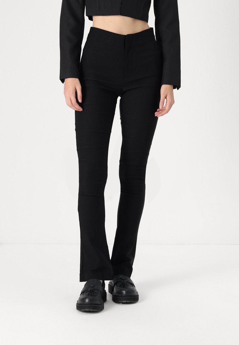 Calças pretas ajustadas com cintura alta, apresentando uma textura suave e costuras mínimas, acompanhadas de sapatos pretos. Cor sólida, sem padrões.