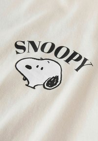 Kreemjas puuvillane T-särk musta väljakujunenud Snoopy graafikaga, kus on kujutatud tegelase pead ja nimi "SNOOPY" julgelt kirjutatud fondis.
