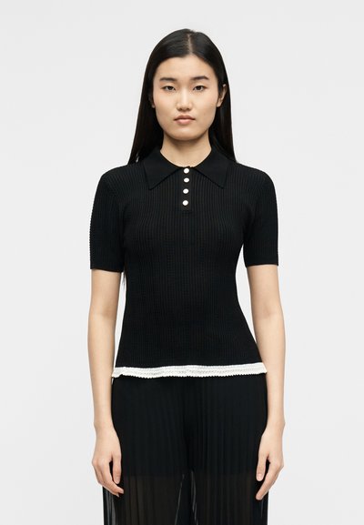 Γυναίκα με μακριά μαύρα μαλλιά φοράει μαύρο ριμπ κοντομάνικο τοπ με λευκά κουμπιά και δαντέλα στο τελείωμα, συνδυασμένο με διάφανο μαύρο πλισέ παντελόνι.