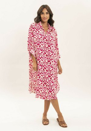 Femme portant une robe ample mi-longue avec un motif abstrait rose et blanc, et des sandales slides marron, debout sur un fond uni.