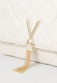 Prošívaná bílá kabelka se zlatým kování; má logo ve tvaru „V“ a detail s řetízkovým třásněm. Hladký povrch s jemným finišem.