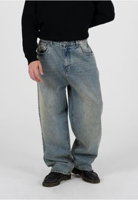 Vaqueros de mezclilla oversize en azul claro con acabado desgastado, diseño de cinco bolsillos y cierre de botones, combinados con zapatos de cuero negros.