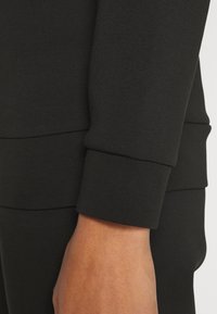 Černá mikina s přiléhavým střihem, ribbenými manžetami, hladkým povrchem a kontrastním švem podél rukávu.