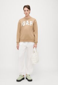 Sweatshirt cáqui com impressão floral "DAD", calças brancas de perna larga, sapatos verde-claro e uma pequena bolsa branca franzida.