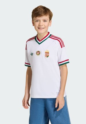 Junge trägt weißes Fußballtrikot mit rotem und grünem Rand sowie ungarischen Wappen, kombiniert mit blauen Jeanshosen, lächelnd und stehend.