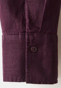 Poignet de chemise en lin violet foncé avec un seul bouton assorti et un tissu tissé texturé visible.
