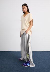 Béžový oversize pletený svetr s výstřihem do V, v kombinaci se stříbrnými saténovými širokými kalhotami s černými pruhy a cvočky a fialovými sportovními botami.