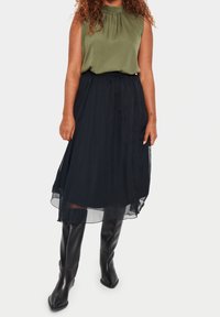 Sleeveless olive green top with gathered neckline paired with a black, flowing, sheer midi skirt and knee-high black leather boots.