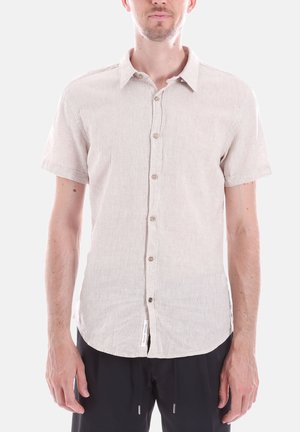 Uomo con una camicia beige chiaro a maniche corte abbottonata e pantaloni neri, in piedi davanti a uno sfondo chiaro e uniforme.