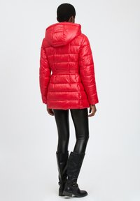 Chaqueta de plumas roja con capucha, diseño acolchado, cintura ajustada y textura brillante. Llevada sobre leggins de cuero negro y botas altas negras.