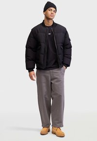 Giacca imbottita nera con cerniera, indossata su un maglione nero, abbinata a pantaloni grigi e stivaletti color cuoio. Il modello indossa anche un berretto nero.