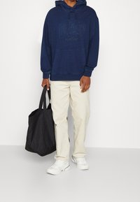 Marineblaue Kapuzenjacke aus strukturiertem Stoff, mit "DAP"-Logo, Kängurutasche, kombiniert mit beigen Hosen und einer schwarzen Tote-Tasche.