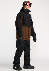 Sneeuwboardjack in zwart en bruin, met een gestructureerd ontwerp. Gecombineerd met zwarte broek, handschoenen en zwarte schoenen, met witte accenten.