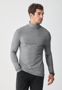 Camisa gris de cuello alto y mangas largas hecha de un tejido suave y elástico. Presenta un diseño ajustado y un cuello alto. Se lleva con jeans oscuros.