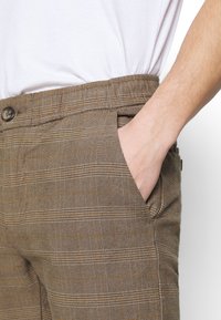 Pantalons écossais marron en tissu texturé, présentant une fermeture à bouton et des poches latérales, mettant en valeur un motif à carreaux avec des accents orange.